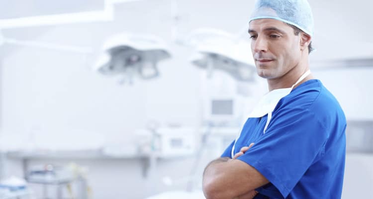 Imagem de médico usando uniforme azul, pensativo e de braços cruzados